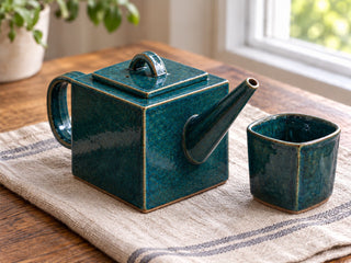 A square slab-built stoneware teapot in glossy teal glaze with a straight cone spout, a flat lid and a strap handle, beside a matching cup, made from The Clay Hole paper template
