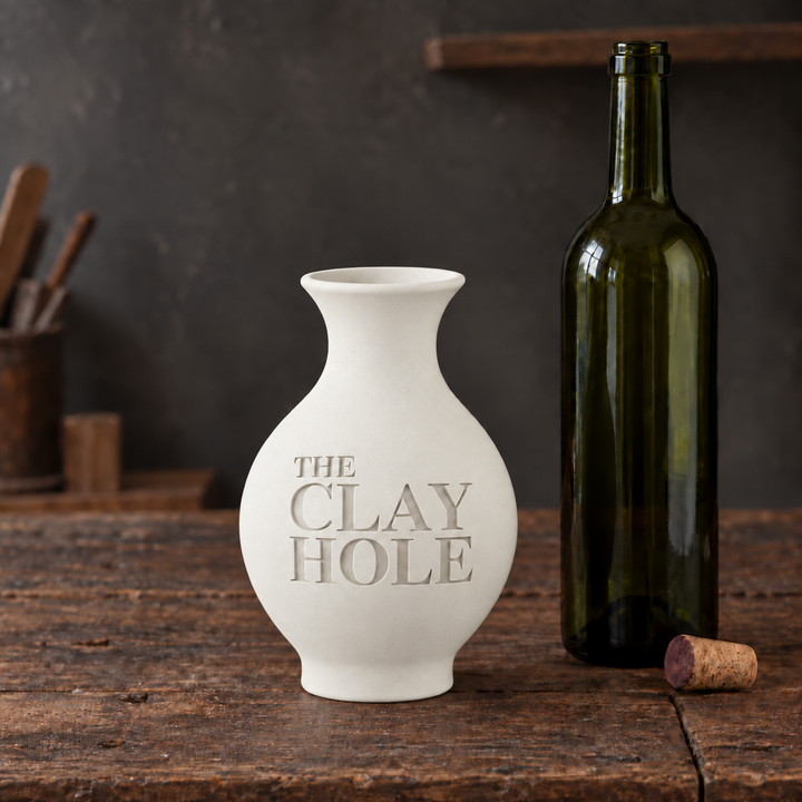 Unglazed white Laguna #16 Porcelain cone 6 clay vase stamped The Clay Hole, beside a wine bottle on a wood table