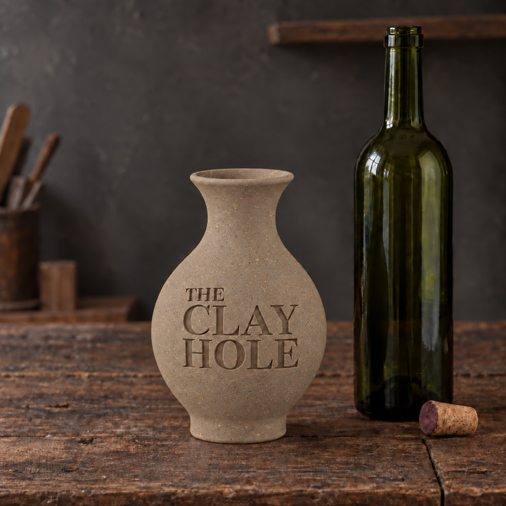 Unglazed tan Laguna 52 Buff clay vase stamped The Clay Hole, beside a wine bottle on a wood table