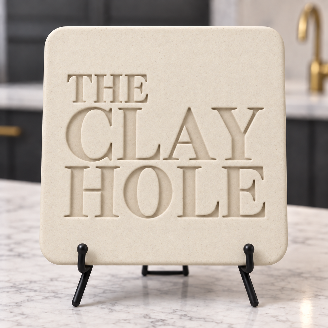 Laguna B-Mix clay tile in cream, stamped with The Clay Hole logo, on a black easel on a marble counter