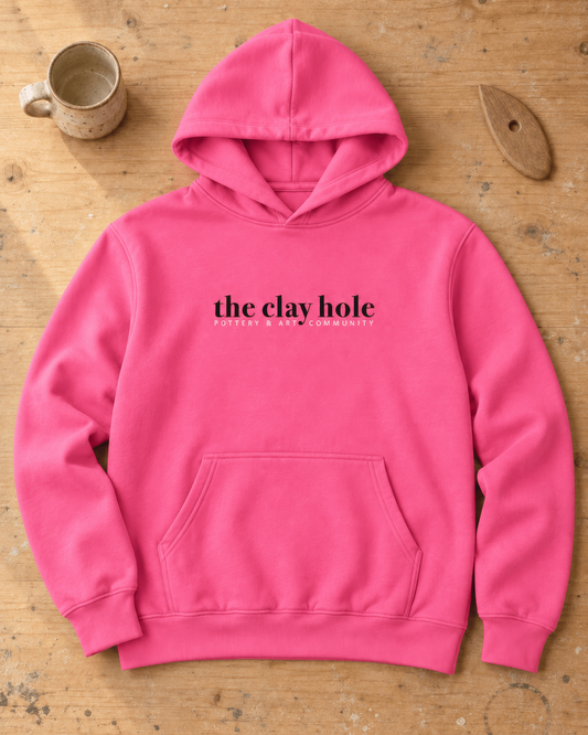 Hot pink The Clay Hole hoodie without drawstrings laid flat on a wooden studio table