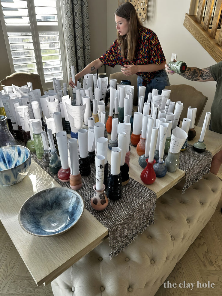 Small glazed bud vases each holding a rolled order slip during a pottery drop