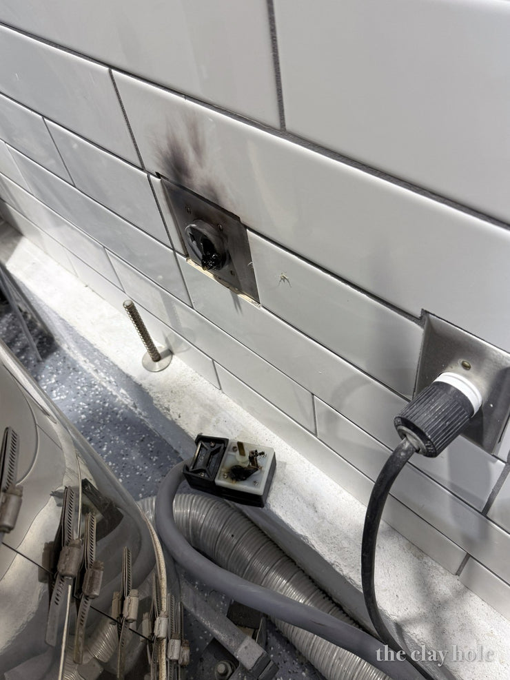 Scorched wall outlet and melted kiln plug on white tile behind a pottery kiln