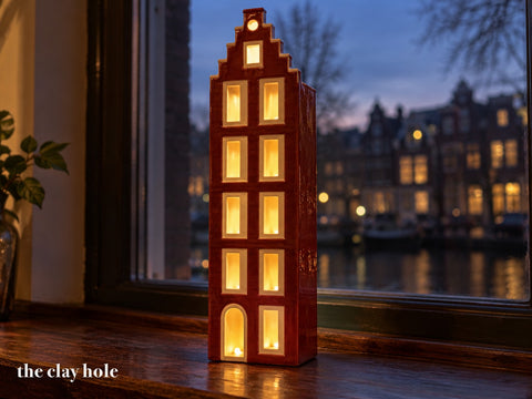 A tall narrow stoneware canal house lantern made from The Clay Hole paper template, every window glowing