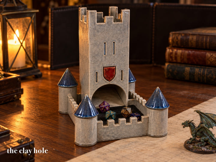 A stoneware castle dice tower with a tall keep, a felt dice courtyard and four blue-roofed towers, made from The Clay Hole paper template