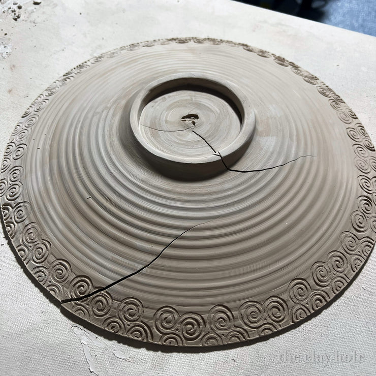 Underside of a leather hard pottery platter with a stamped spiral rim and long drying cracks