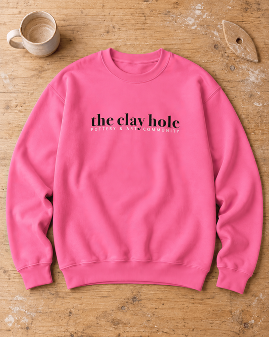 Pink The Clay Hole crewneck sweatshirt laid flat on a wooden studio table