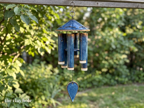 A handmade stoneware wind chime with six blue glazed tubes under a cone roof, a round striker and a teardrop sail, made from a Clay Hole paper template