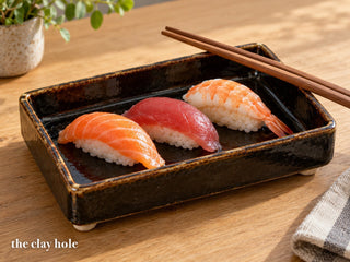 A folded black stoneware serving tray holding nigiri sushi, made from The Clay Hole folded tray paper template