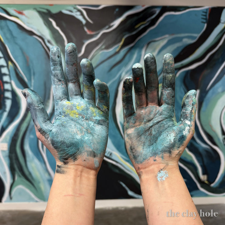 Two hands covered in blue glaze held up in front of the mural at The Clay Hole pottery studio