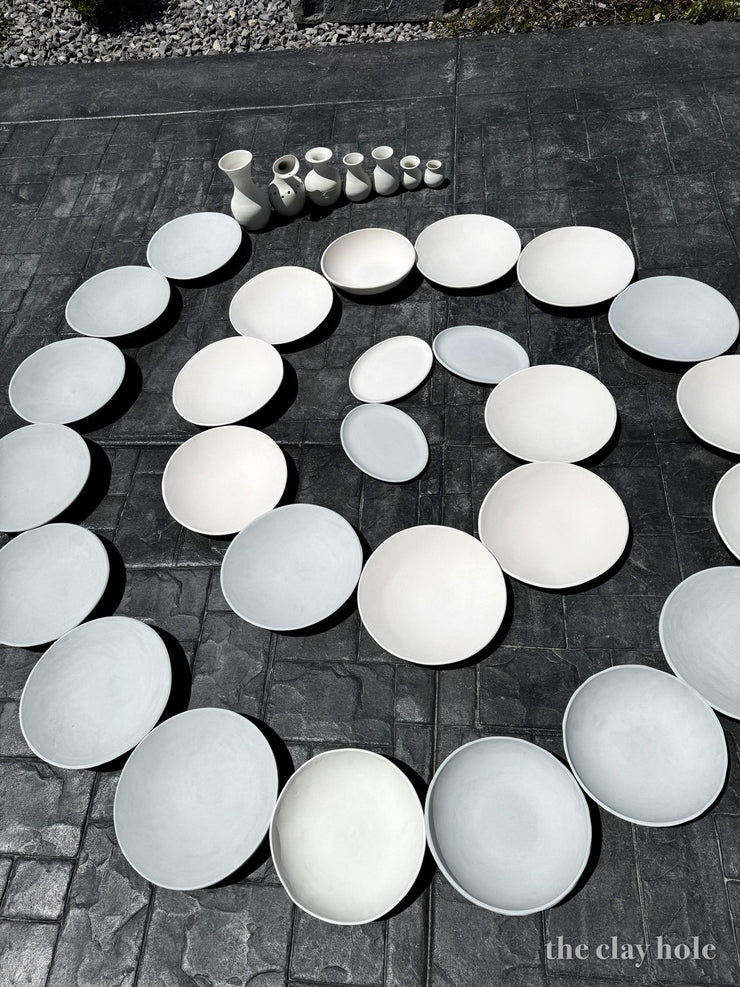 Unfired plates and bowls arranged in a spiral on stamped concrete, drying in the sun