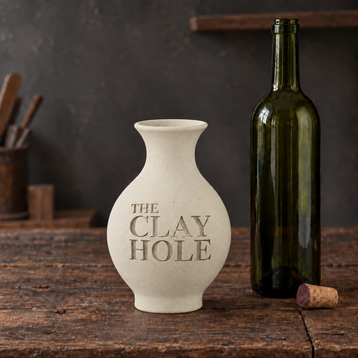 Unglazed off-white Laguna Half and Half clay vase stamped The Clay Hole, beside a wine bottle on a wood table