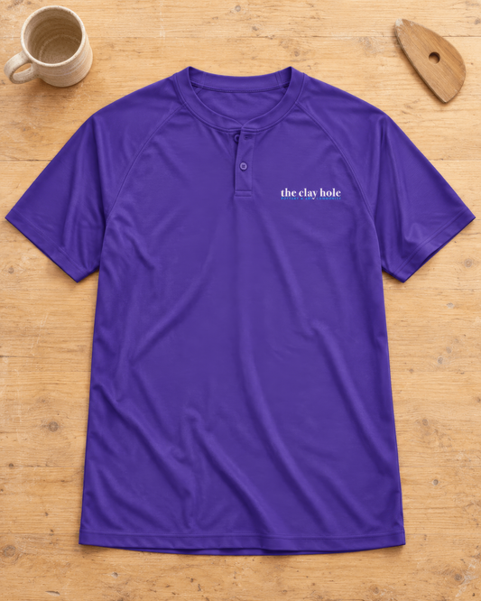 Purple The Clay Hole men's henley jersey shirt laid flat on a wooden studio table