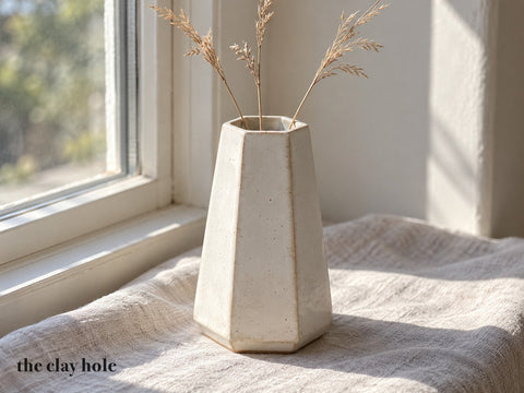A six-sided white stoneware bud vase with mitered corners holding dried grass, made from The Clay Hole hexagon vase paper template