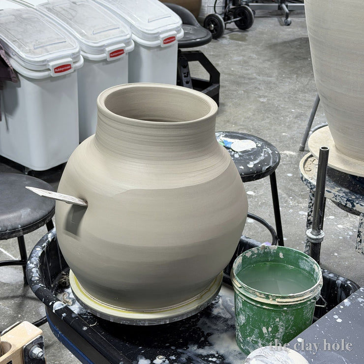 Large freshly thrown pot in wet clay on the wheel with a wooden tool resting in its side