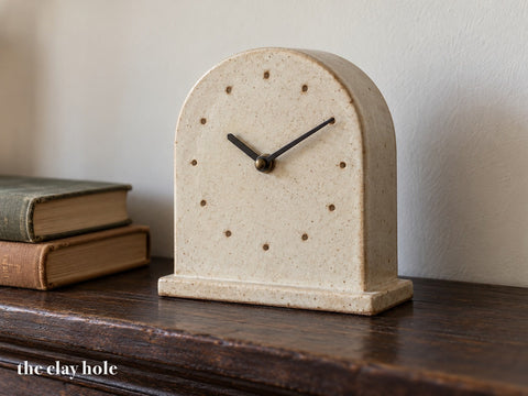 An arch-topped slab-built stoneware mantel clock in matte oatmeal glaze with twelve pressed hour dots, standing on a wooden mantel, made from The Clay Hole paper template