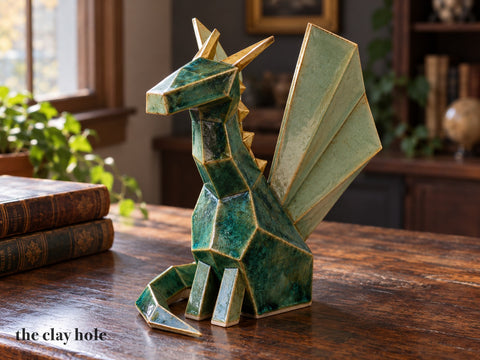 A handmade stoneware origami dragon in jade green with celadon wings and gold horns, built from flat faceted slab pieces with a Clay Hole paper template