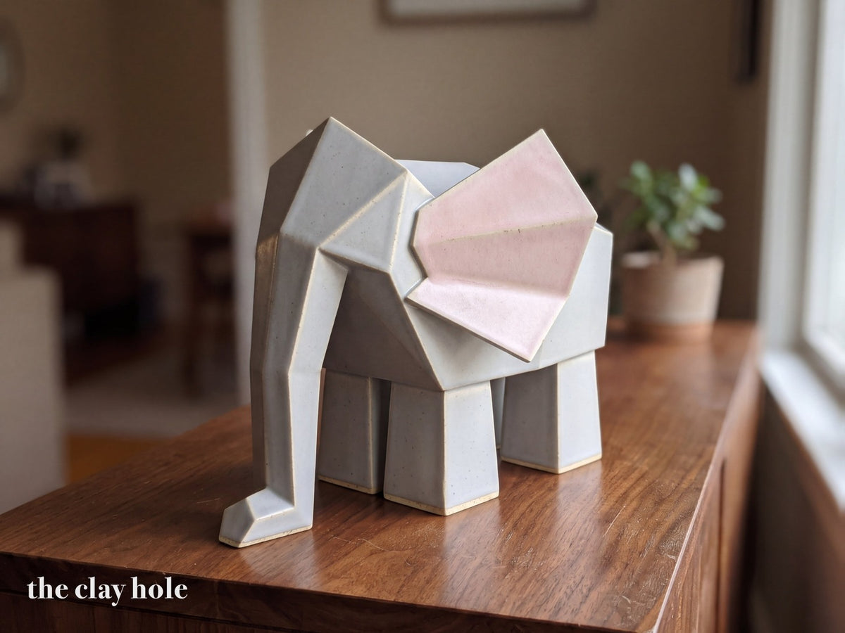 A handmade stoneware origami elephant in dove gray with pink ears, built from flat faceted slab pieces with a Clay Hole paper template