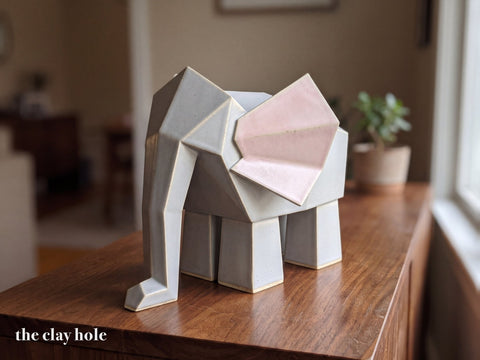 A handmade stoneware origami elephant in dove gray with pink ears, built from flat faceted slab pieces with a Clay Hole paper template