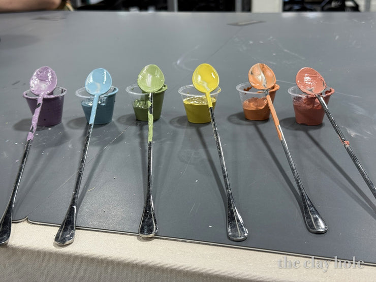 Six spoons dripping pastel pottery glaze samples resting across small sample cups