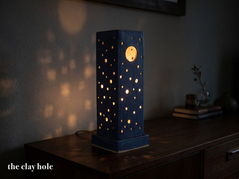A tall square pierced stoneware lantern lamp made from The Clay Hole paper template, glowing with dots of light