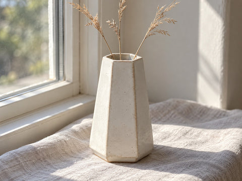 A six-sided white stoneware bud vase with mitered corners holding dried grass, made from The Clay Hole hexagon vase paper template