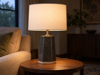 A six-sided slab-built stoneware table lamp body in speckled charcoal glaze with a white drum shade, lit, on a side table, made from The Clay Hole paper template