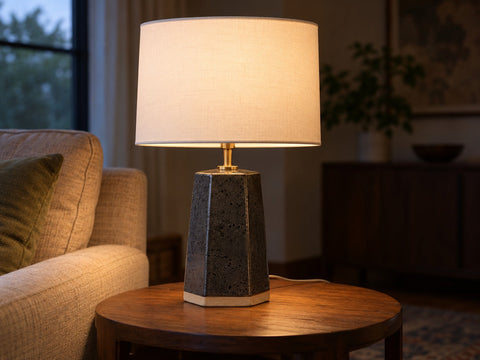 A six-sided slab-built stoneware table lamp body in speckled charcoal glaze with a white drum shade, lit, on a side table, made from The Clay Hole paper template