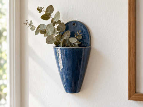 A deep blue stoneware wall pocket vase hanging on a wall with eucalyptus, made from The Clay Hole wall pocket paper template