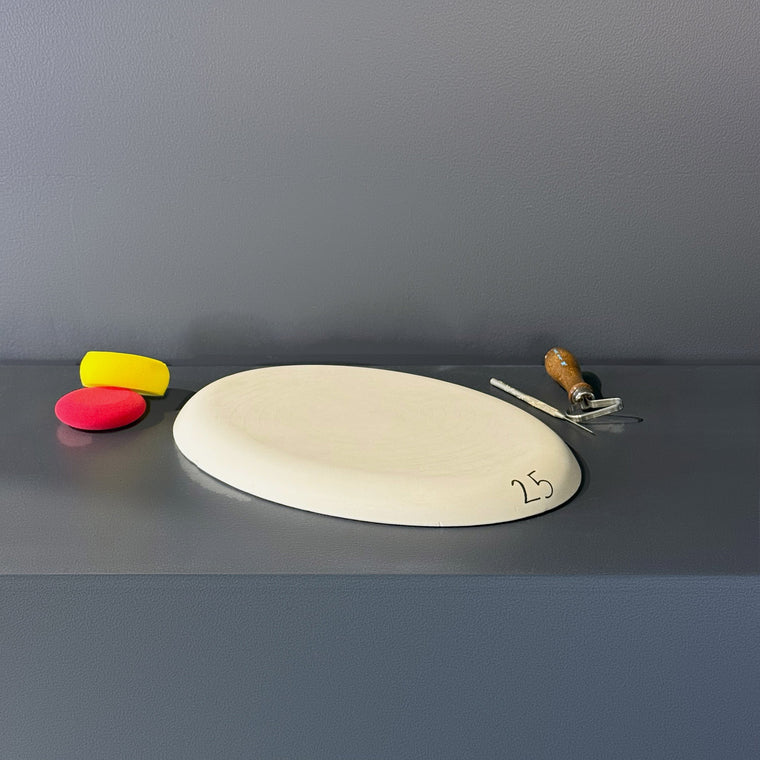 Flat white plaster oval slab mold number 25: a nearly flat oval, measuring 14.62 in wide, 10.38 in deep and 1 in tall and an inch thick, lying on the gray shelf with a hand-written 25 on the surface.