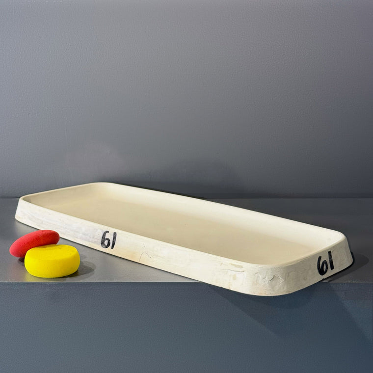 Large white plaster slump mold number 61: an elongated rectangular tray mold, measuring 22 in wide, 9.75 in deep and 2 in tall with shallow upturned walls, lying concave side up on the gray shelf, hand-numbered 61 at both ends.