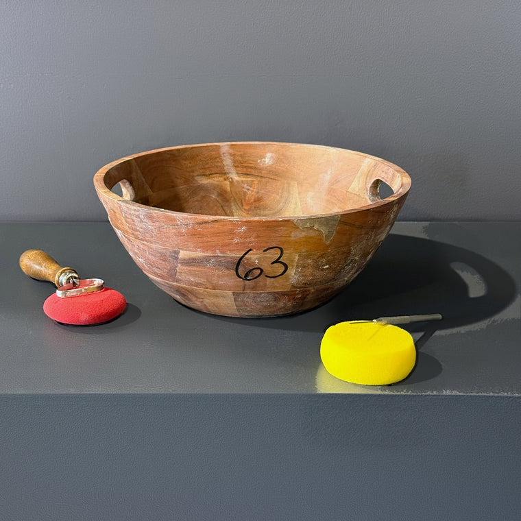 Wooden bowl used as mold number 63: a bowl, measuring 11.4 in wide, 11.4 in deep and 5.2 in tall with two cut-out handle openings in the rim, sitting upright on the gray shelf.