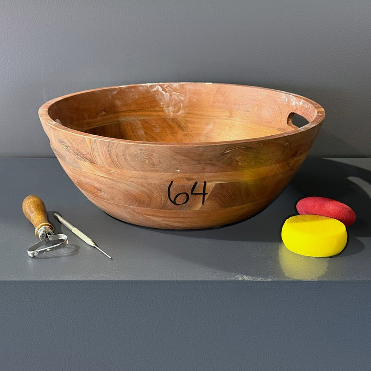 Large wooden bowl used as mold number 64: a bowl, measuring 15 in wide, 15 in deep and 6.6 in tall with two cut-out handle openings, sitting upright on the gray shelf.