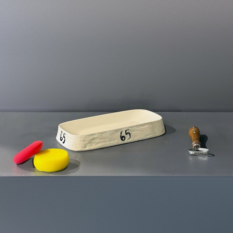 White plaster slump mold number 65: a small elongated tray mold, measuring 10.88 in wide, 5.5 in deep and 1.62 in tall with shallow walls, lying concave side up at an angle on the gray shelf, hand-numbered 65 on both ends.