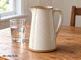 A tall tapered slab-built stoneware pitcher in glossy white glaze with a pinched spout and a strap handle, beside a glass of water, made from The Clay Hole paper template