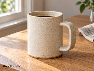 A straight-sided handmade speckled stoneware slab mug with a strip handle, made from The Clay Hole paper template, filled with coffee on a wooden table