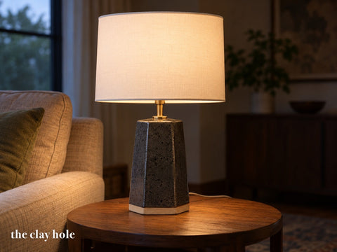A six-sided slab-built stoneware table lamp body in speckled charcoal glaze with a white drum shade, lit, on a side table, made from The Clay Hole paper template
