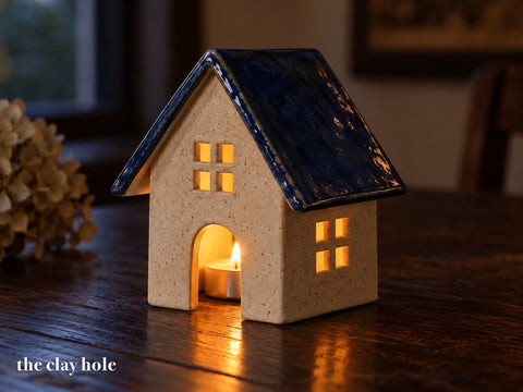 A small stoneware tealight house with a blue roof glowing from a candle inside, made from The Clay Hole tealight house paper template