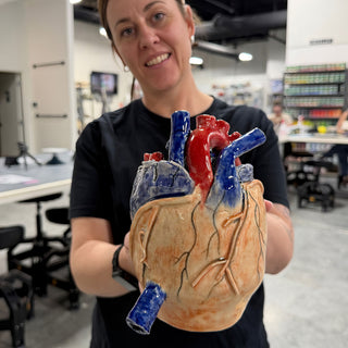 A member holding out a heart sculpture with peach and orange glaze, red and blue vessels and raised veins at The Clay Hole pottery studio in Draper, Utah