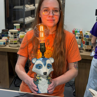 A member holding a lamp shaped like an animal with a white face, blue-gray eye patches and a lit bulb at The Clay Hole pottery studio in Draper, Utah