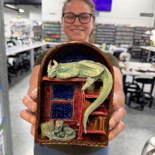 A member holding an arched scene of a green dragon draped over a red bookshelf with a small creature below at The Clay Hole pottery studio in Draper, Utah