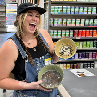 A member winking while holding a cream bowl with a black bird and a green bowl with a spiral inside at The Clay Hole pottery studio in Draper, Utah
