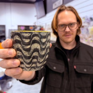 A member holding up a cup with black glaze, cream colored wavy lines and a yellow inside rim at The Clay Hole pottery studio in Draper, Utah
