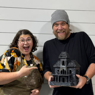 Two members posing with a black sculpted haunted house with a shingled roof, tall windows and a front porch at The Clay Hole pottery studio in Draper, Utah