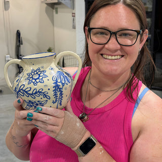 A member holding a cream two-handled vase painted with blue flowers and leaves and a blue rim at The Clay Hole pottery studio in Draper, Utah