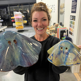 A member holding two wide wing shaped sculptures with two round holes each, one blue and one yellow at The Clay Hole pottery studio in Draper, Utah