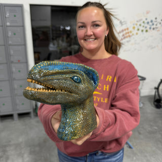 A member holding a dinosaur head with scaly blue and green glaze, an open mouth and visible teeth at The Clay Hole pottery studio in Draper, Utah