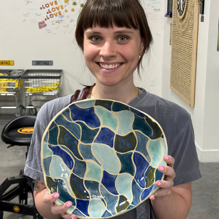 A member holding a wide plate with blue and teal wavy shapes separated by thin cream lines at The Clay Hole pottery studio in Draper, Utah
