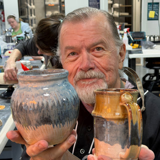 A member holding a rounded vase with blue, gray and peach drips and a tall mug in amber and brown at The Clay Hole pottery studio in Draper, Utah