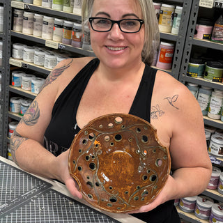 A member holding a round amber brown bowl with scattered round cutout holes and carved vine lines at The Clay Hole pottery studio in Draper, Utah